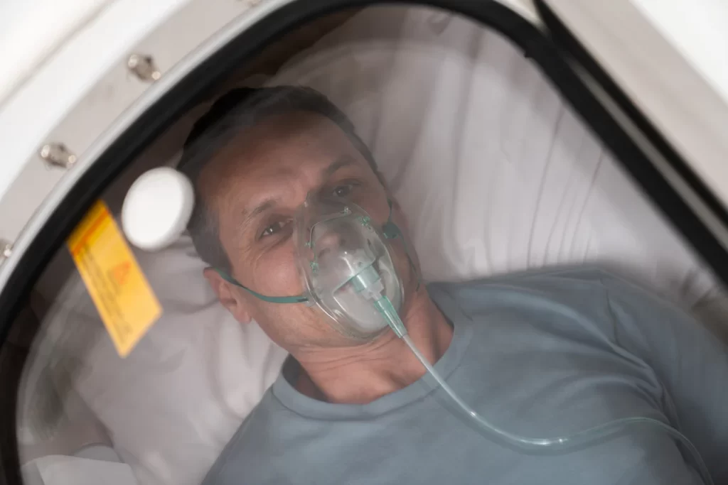 A person in a hardshell hyperbaric oxygen therapy chamber breathes pure oxygen during a therapy session.